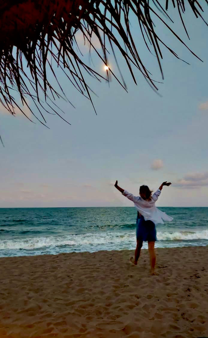 Person standing on a beach with arms outstretched, overlooking the ocean at sunset.